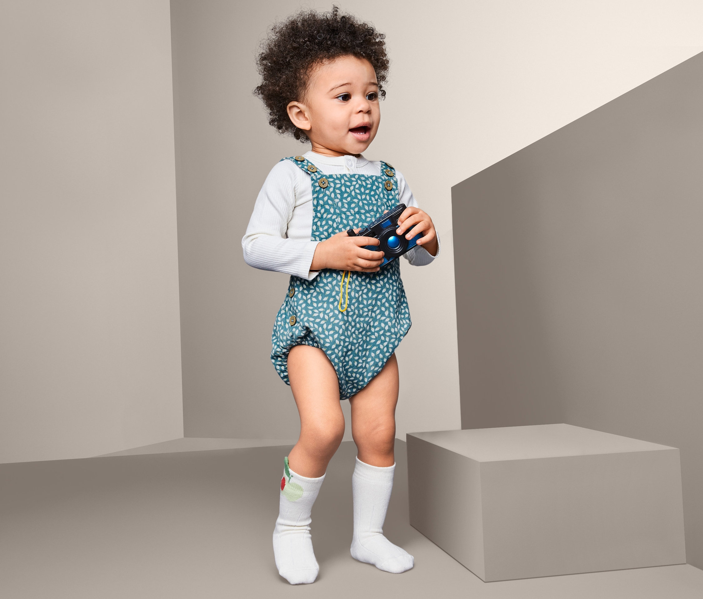 Egy göndör hajú kisgyerek áll, játék fényképezőgépet tart a kezében, és egy kék, leveles kisgyerek napozót visel.