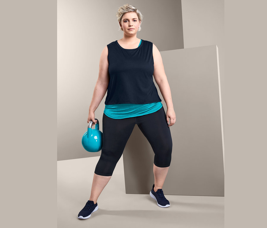 Nő kettlebellt tart, és Női 2 az 1-ben sporttrikót, türkiz/sötétkék és Női 3/4-es sportnadrágot, sötétkéket visel.