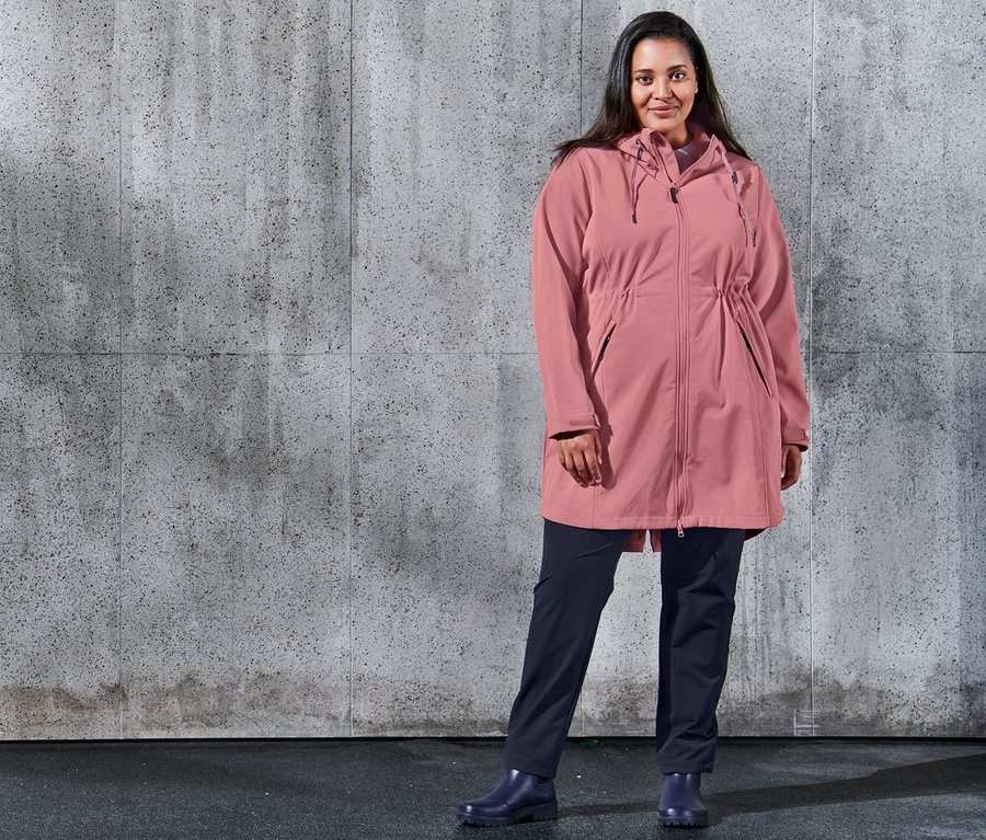 Egy nő pózol egy betonfal előtt, softshell kabátot és sötétkék softshell szabadidőnadrágot visel.
