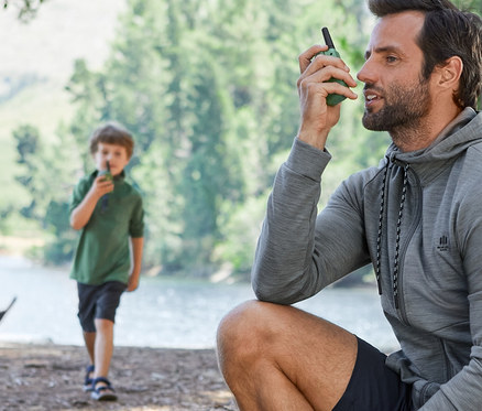 Egy férfi ül egy fatörzsön és walkie-talkie-t használ. A háttérben egy fiú látható walkie-talkie-val. Mindketten funkcionális ruházatot viselnek.