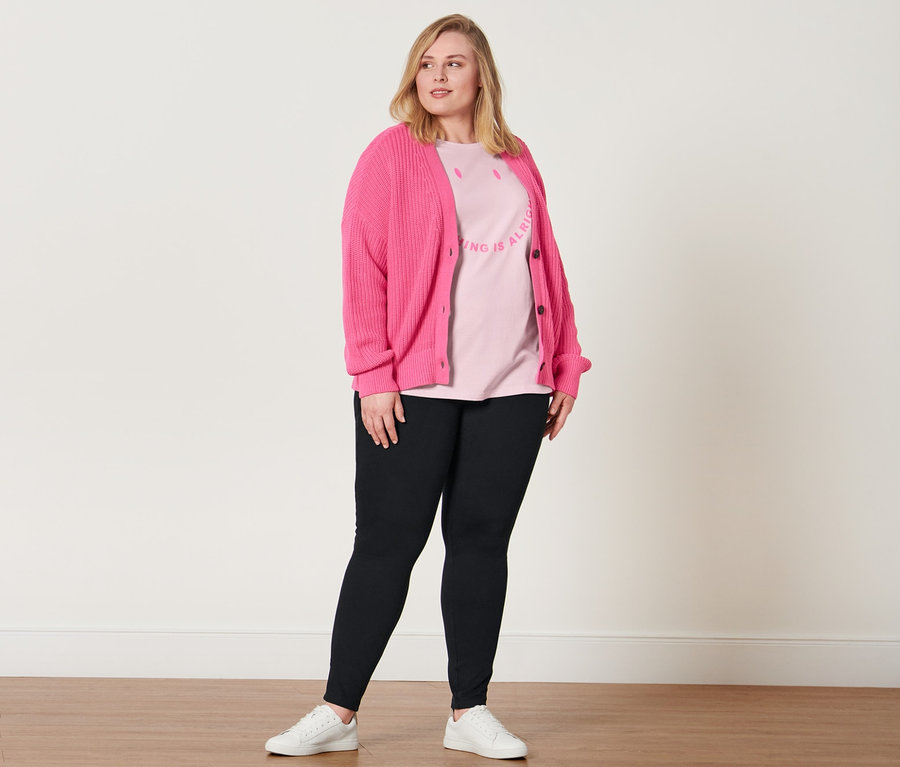 Nő pózol fekete jeggingsben, rózsaszín feliratos pólóban, fehér bőr sneaker cipőben és pink kötött kardigánban.