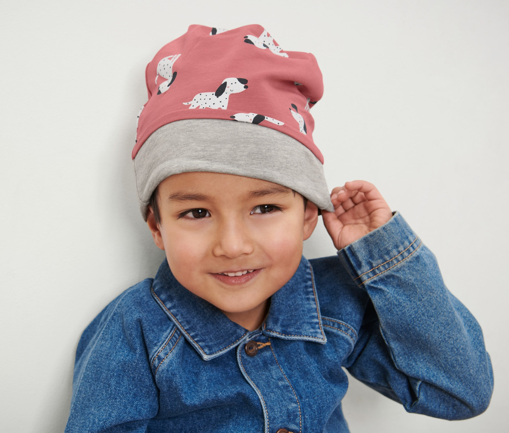 Egy fiú rózsaszín-szürke kifordítható gyereksapkát és farmerkabátot visel.
