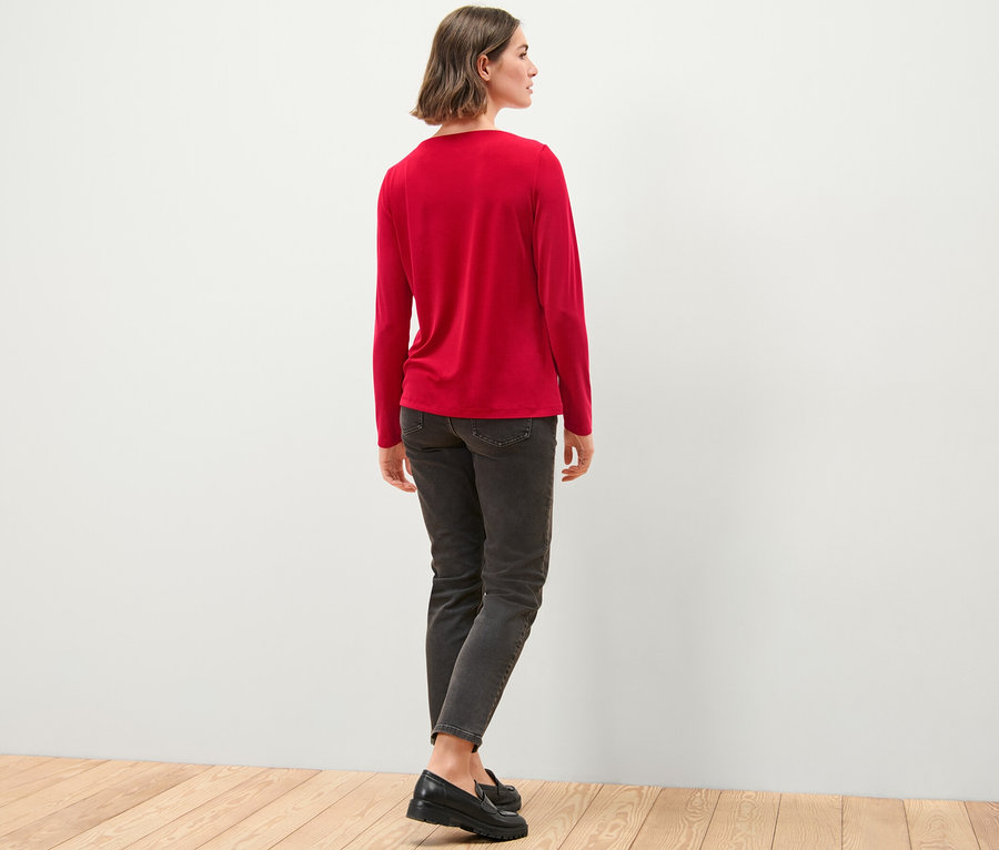 Egy nő áll háttal, és egy piros női hosszú ujjú pólóblúzt, farmert és cipőt visel.