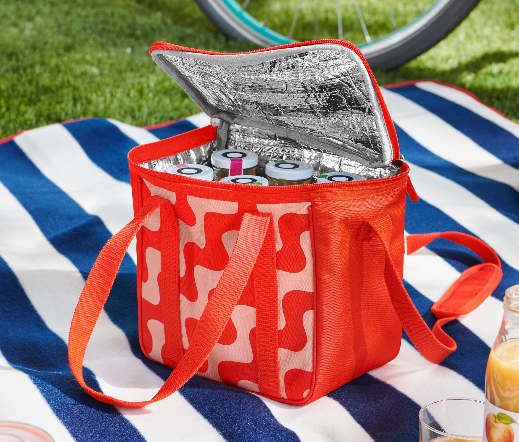 Egy nyitott, piros-fehér mintás hűtőtáska áll egy kék-fehér csíkos takarón, tele üvegekkel. A háttérben egy kerékpár és egy sisak látható.