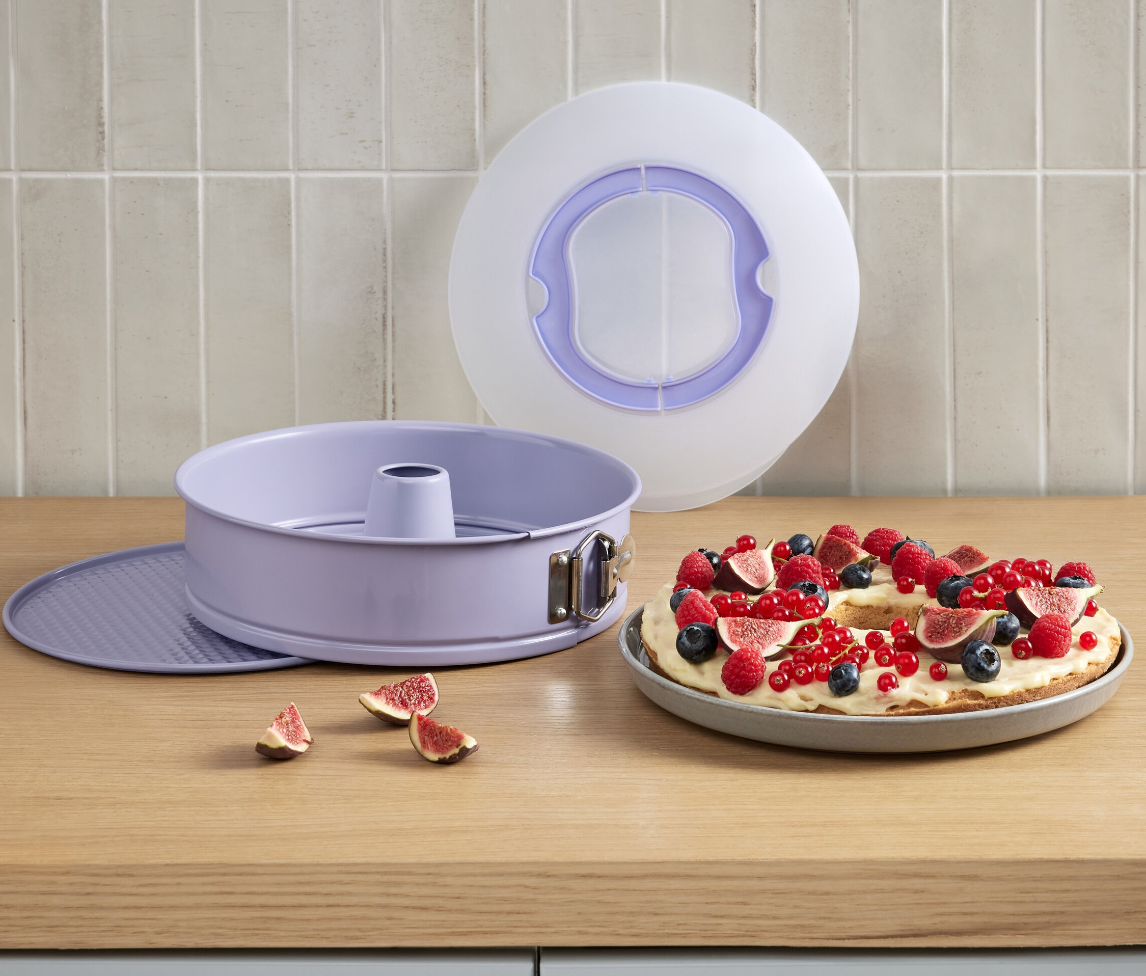 Egy lila tortaforma szállítófüllel egy gyümölcsös sütemény mellett.