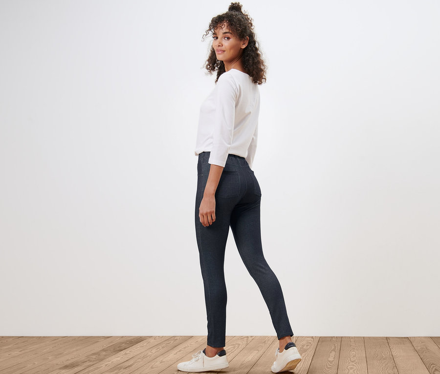 Egy nő visszanéz a válla fölött, farmer hatású leggingset, fehér blúzt és tornacipőt visel.