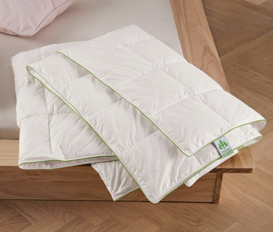 Két párna és egy összehajtott irisette® greenline nyári pehelypaplan normál méretben egy ágyon.