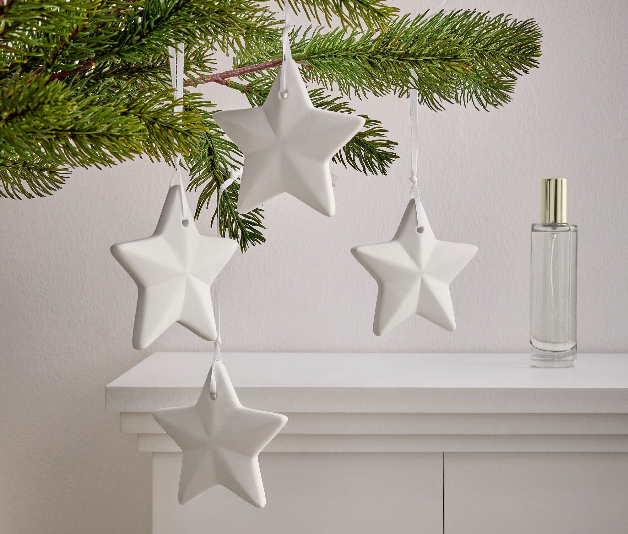 Egy ág tűlevelekkel, amelyen négy fehér csillag lóg egy fehér szekrényen. A csillagok mellett egy parfümös üveg áll.