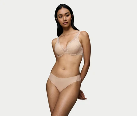 Sötét hajú nő visel egy nude bézs Triumph Body Makeup Illusion WP csipkemelltartót virágos csipkerészletekkel és hozzá illő alsóneműt. Kezei a háta mögött vannak összefonva.