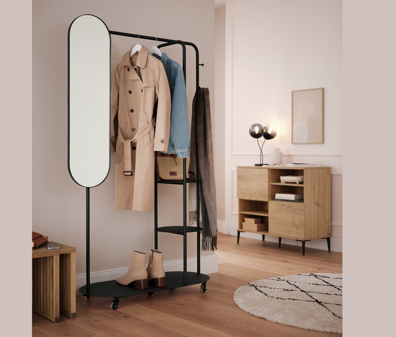 Fekete ruhatartó állvány ovális tükörrel és kihúzható ülőkével. A rudakon bézs kabát, kék dzseki és szürke sál lóg.