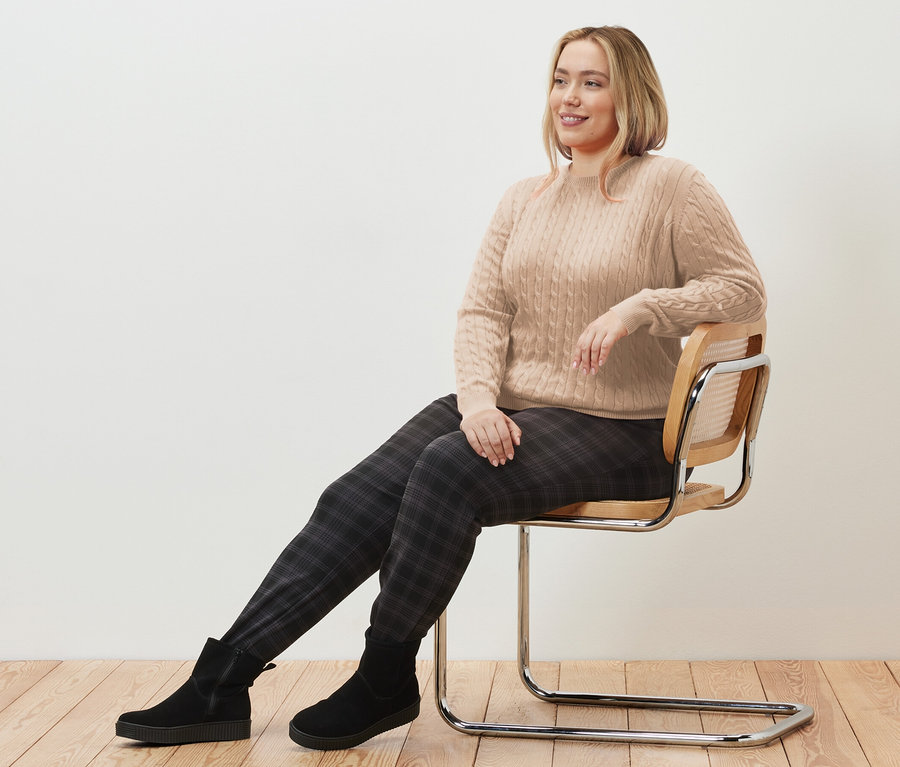 Nő ül egy széken, krémszínű fonott mintás kötött pulóvert és kockás női lezser nadrágot visel.