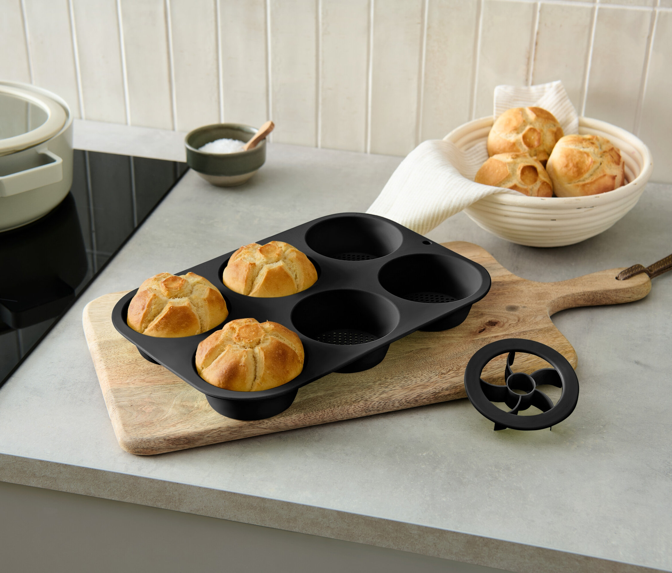 Három sült zsemle egy fekete sütőformában egy fa vágódeszkán egy zsemlés tál mellett.