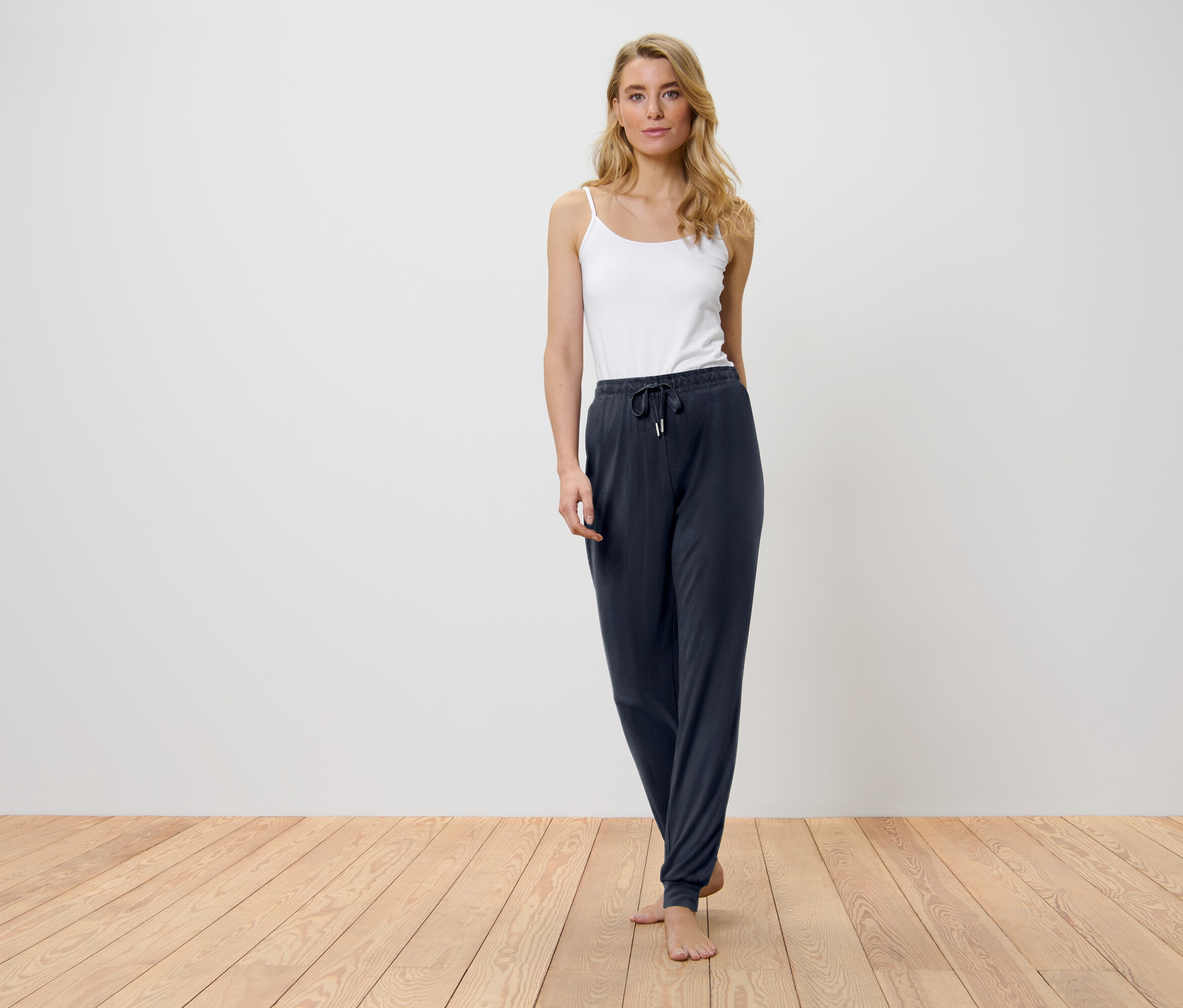 Egy szőke nő áll a fapadlón, fehér felsőben és sötétkék nadrágban.