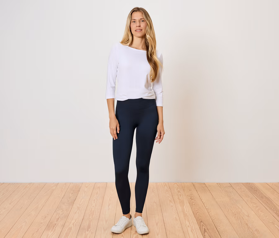 Egy nő áll egy fapadlón, fehér hosszú ujjú inget és fekete leggingset visel.