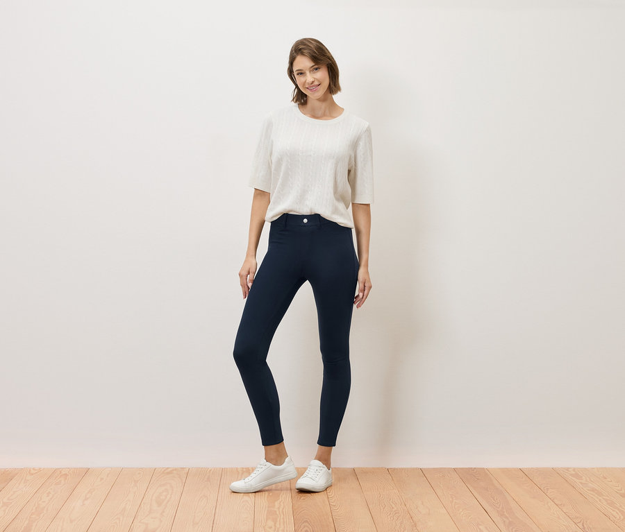 Nő áll krém színű felsőben, sötétkék nadrágban és fehér tornacipőben.