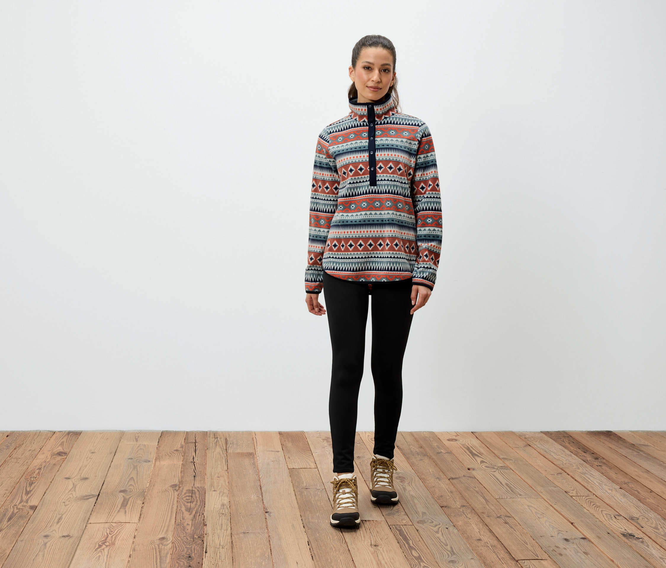 Egy nő áll a fa padlón, mintás pulóvert, fekete leggingset és barna csizmát visel.