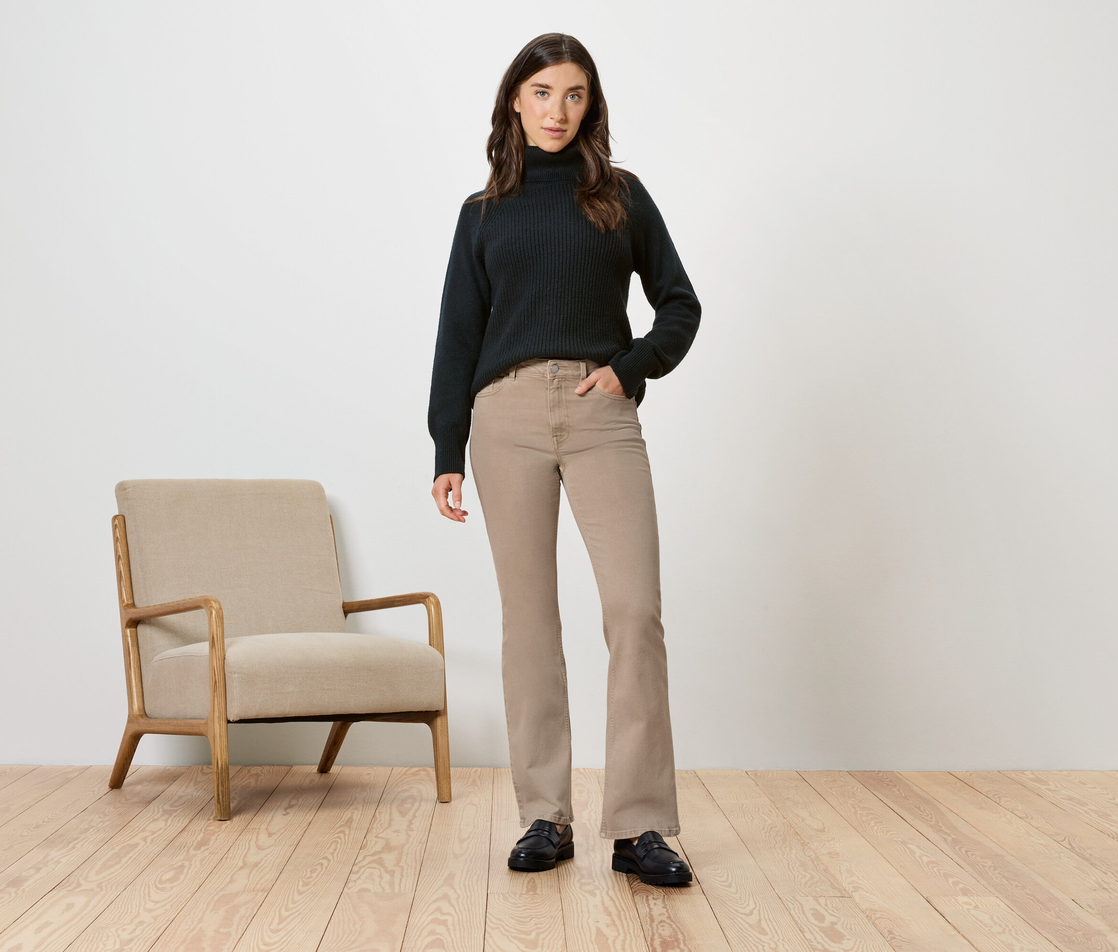 Nő fekete garbóban és bézs nadrágban pózol egy bézs fotel mellett.