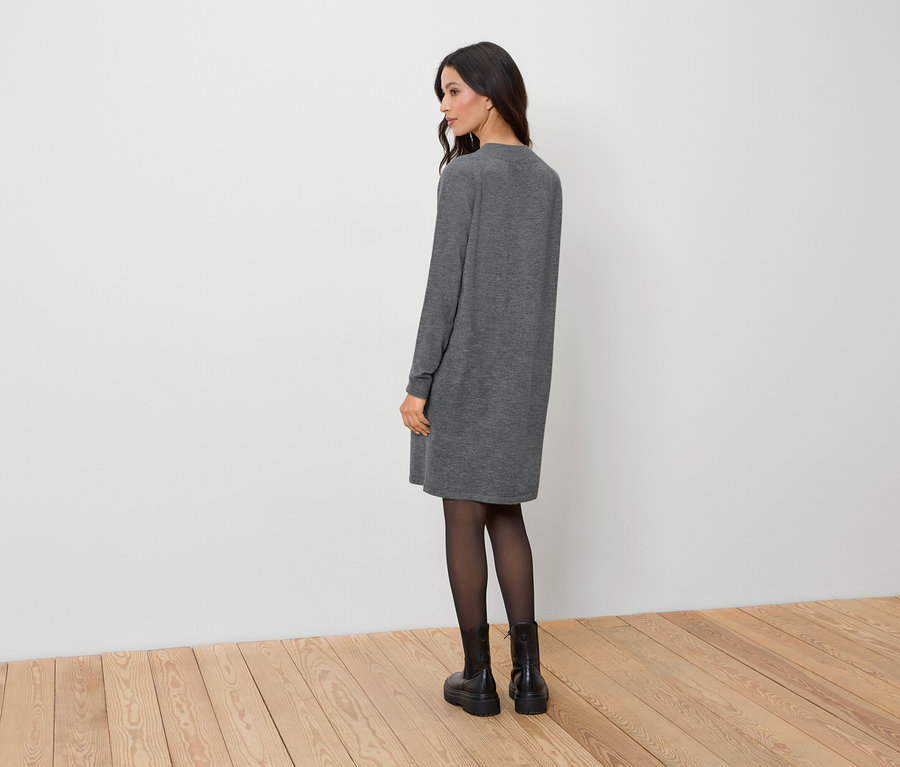 Egy nő háttal áll a kamerának, szürke ruhát, fekete harisnyát és fekete csizmát visel.