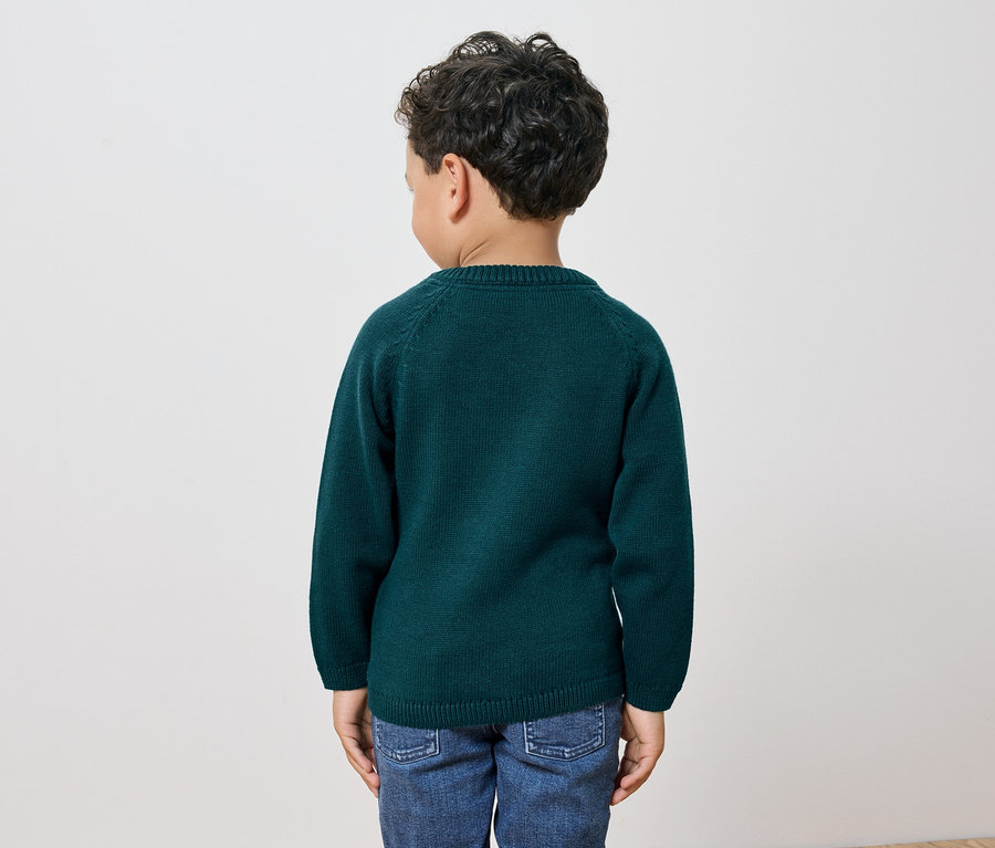 Egy sötét, göndör hajú fiú áll háttal a kamerának, zöld pulóvert és farmert visel.