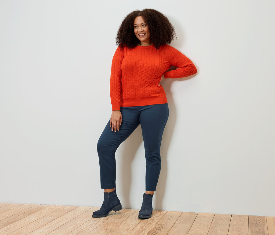 Mosolygó, göndör hajú nő narancssárga pulóverben, kék nadrágban és csizmában pózol.