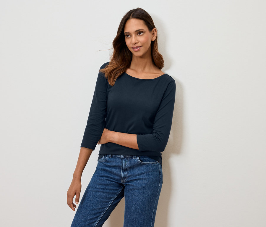 Nő pózol sötétkék háromnegyedes ujjú pólóban és kék farmerben.
