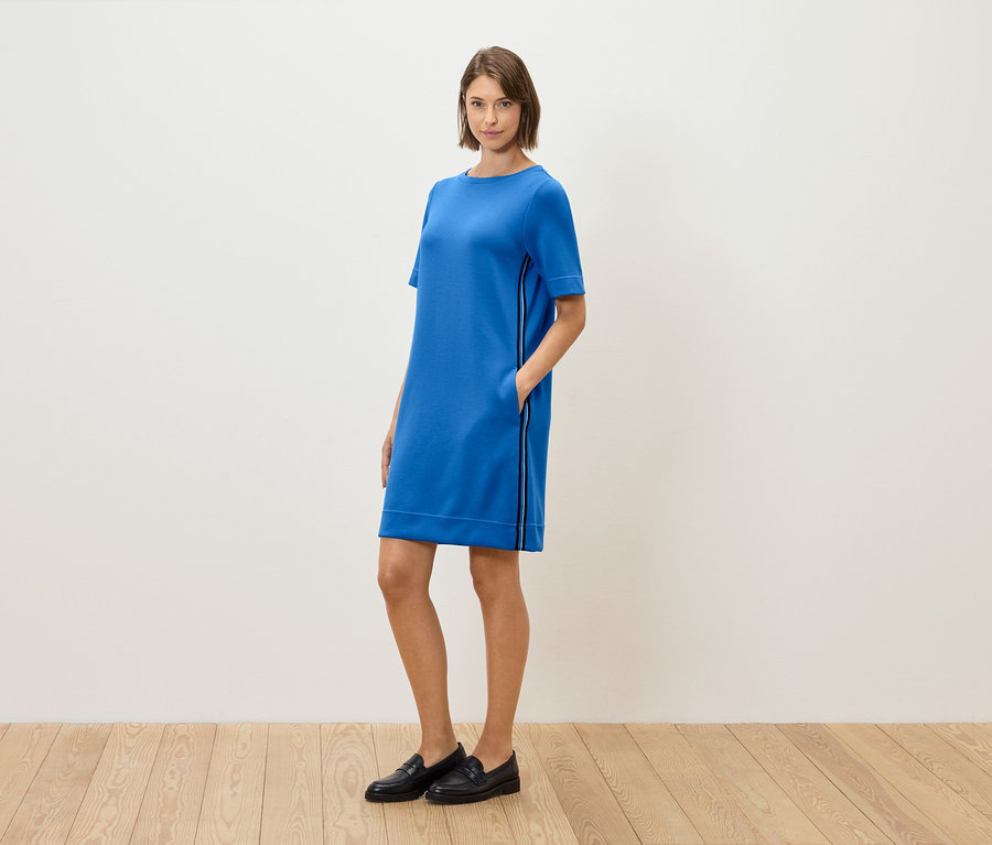 Egy nő áll és pózol egy kék, rövid ujjú ruhában és fekete loaferben.