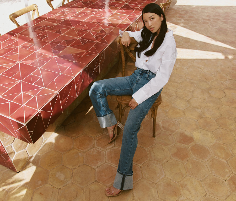 Egy nő ül egy széken egy asztalnál, világoskék Angels »Lara« Straight Fit feltűrt szárú farmert visel.