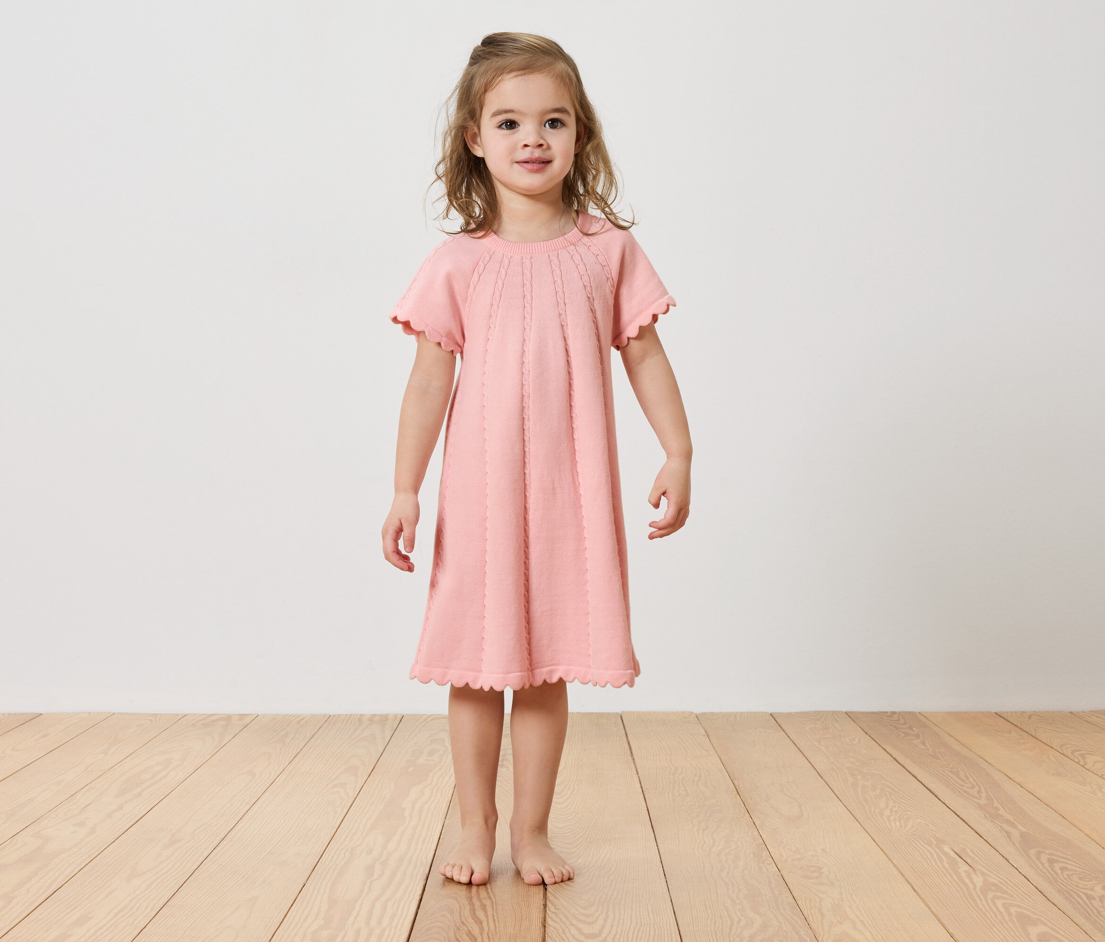 Kislány áll mezítláb a fapadlón, rózsaszín ruhát visel.