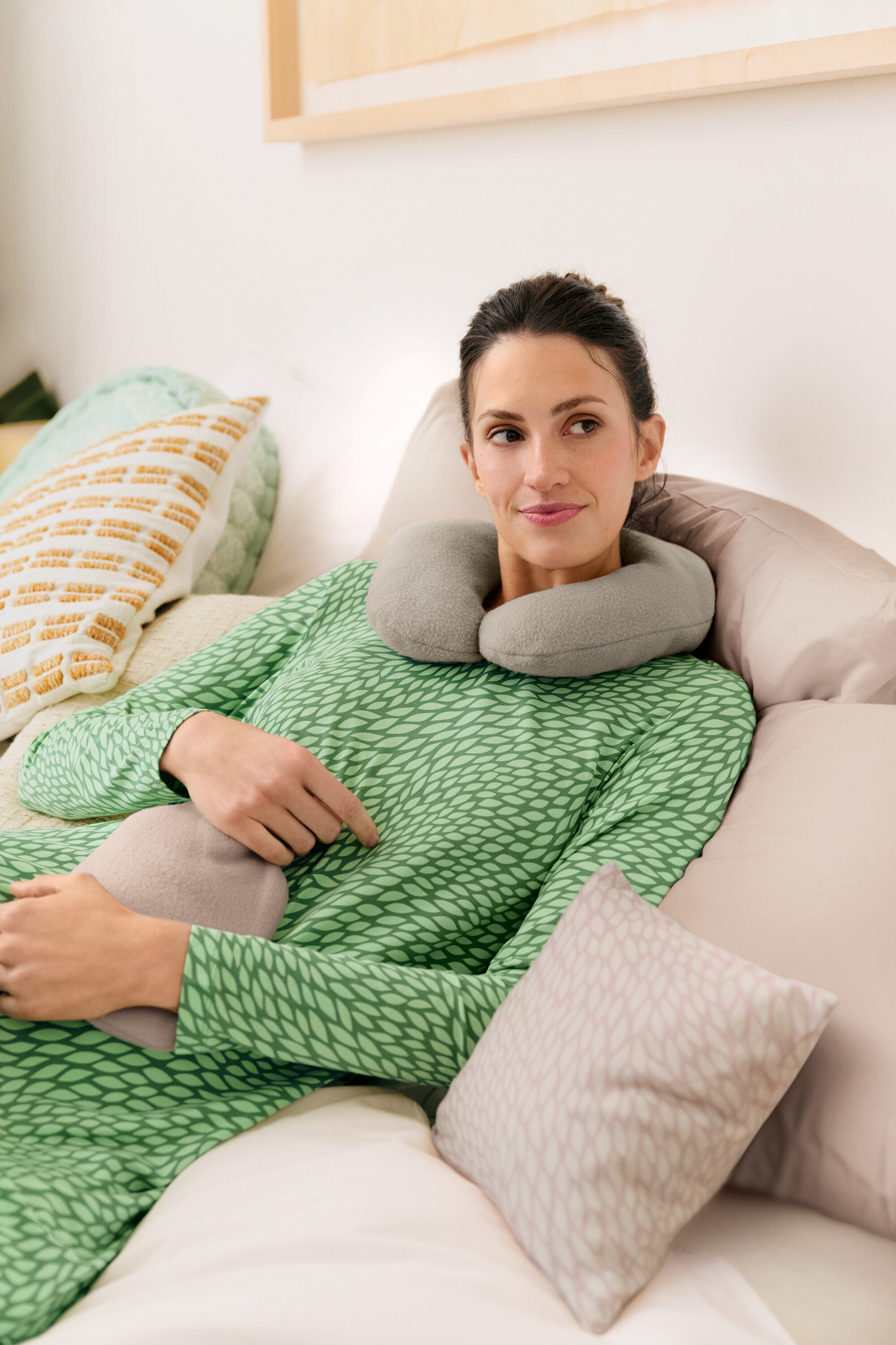 Nő fekszik az ágyban szürke nyakpárnával és hőpalackkal, zöld hálóinget visel.