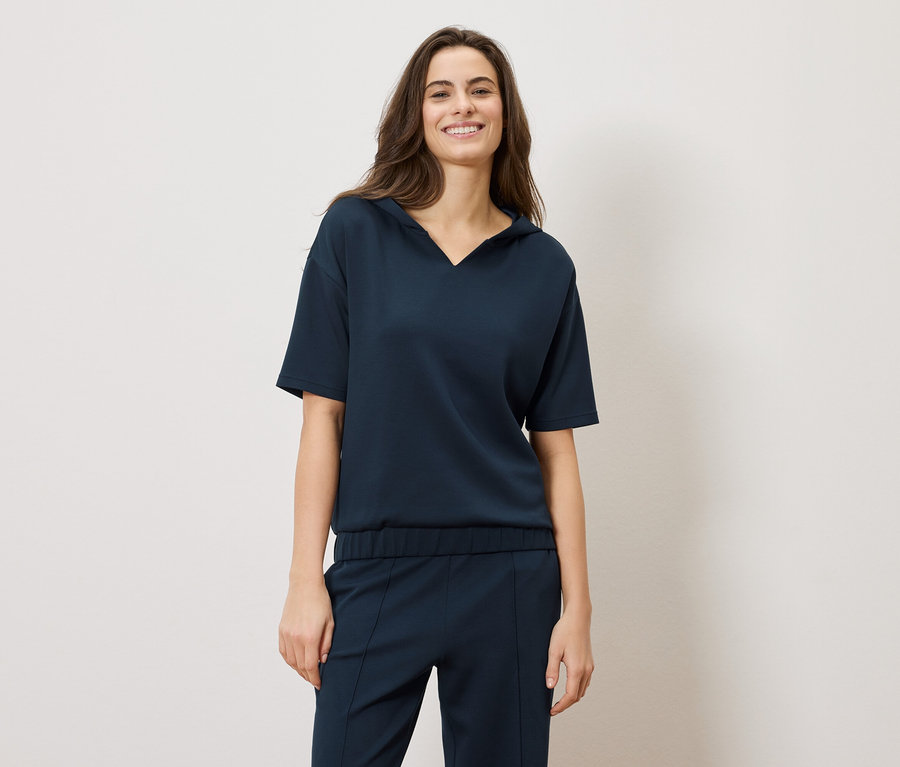 Egy mosolygó, hosszú barna hajú nő sötétkék rövid ujjú felsőt és nadrágot visel.