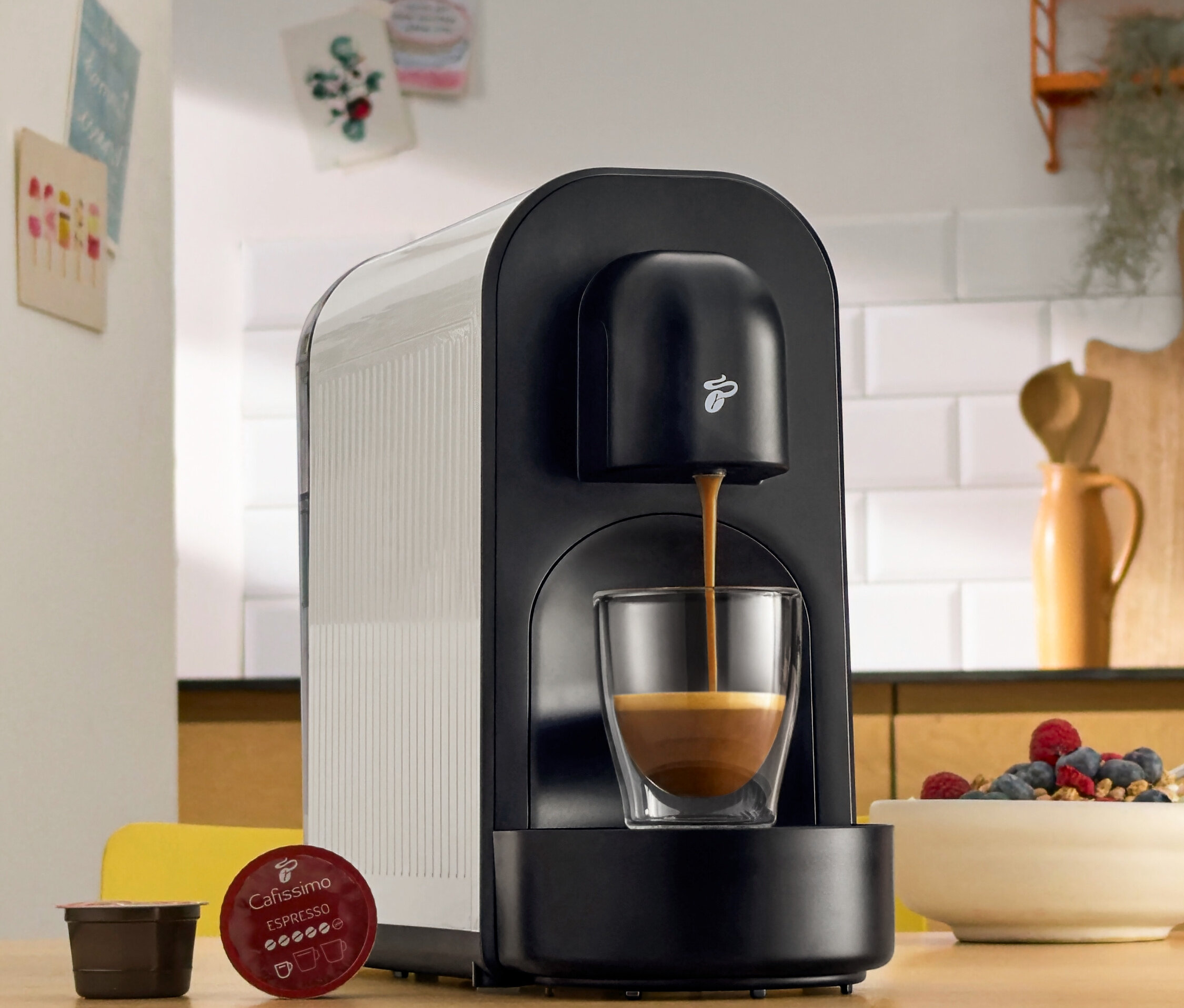 Egy fekete kávéfőző kávét tölt egy pohárba. Mellette kávékapszulák és egy tál bogyós gyümölcs található.