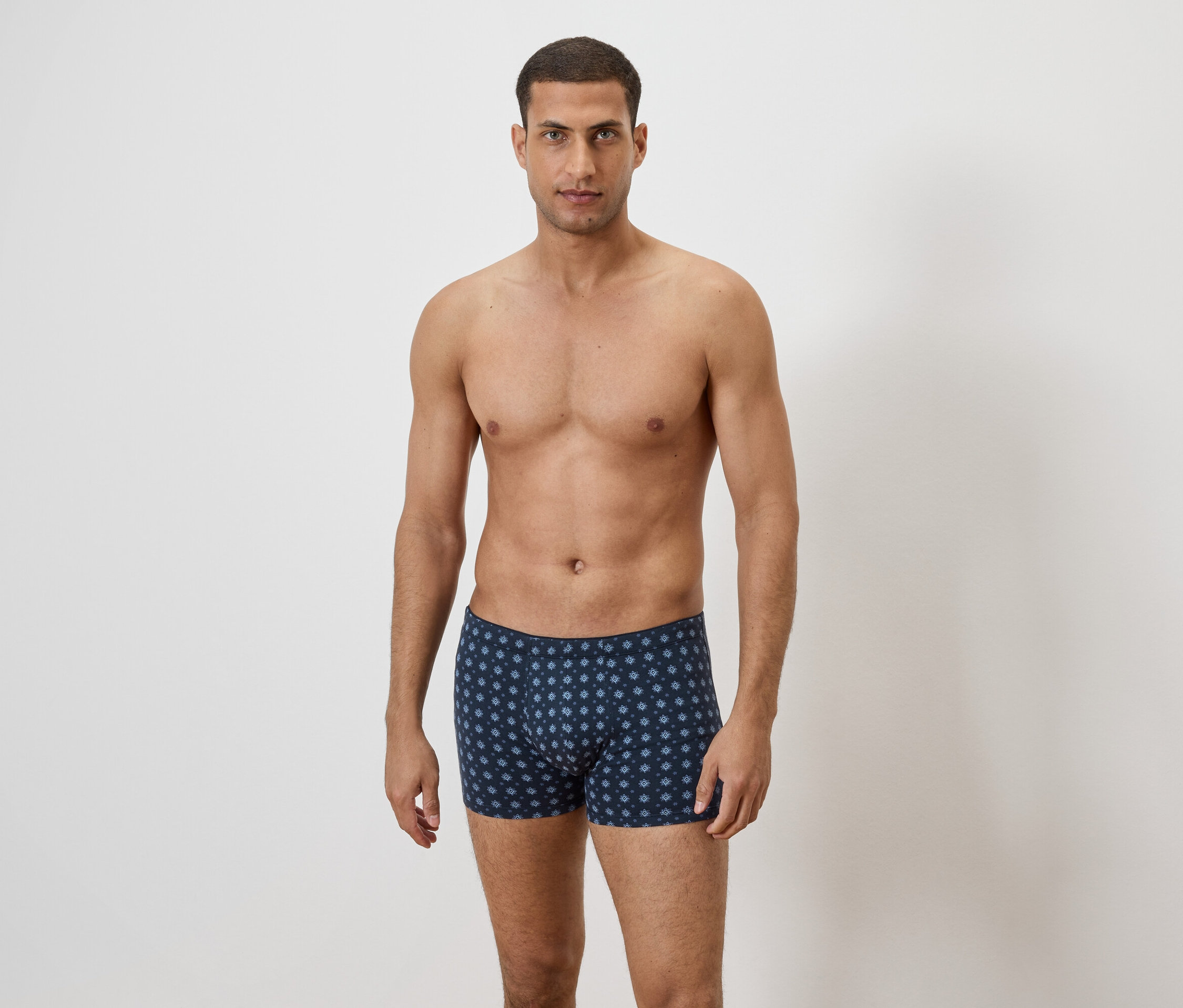 Egy férfi áll meztelen felsőtesttel egy fehér fal előtt, és kék, ismétlődő virágmintás boxeralsót visel.