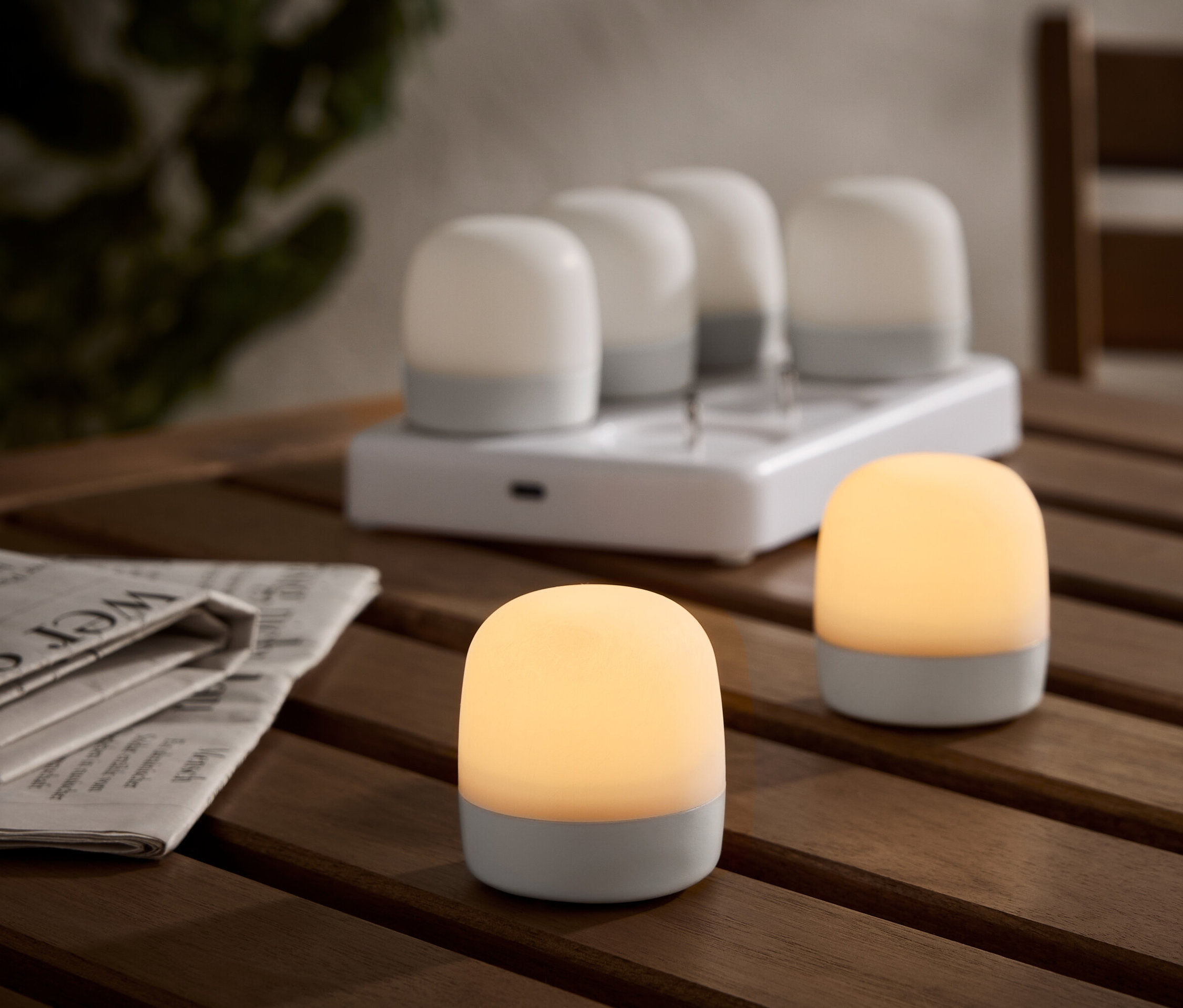 Két világító, vezeték nélküli asztali lámpa egy fa asztalon, töltőállomással és további négy lámpával a háttérben. Mellette egy újság.