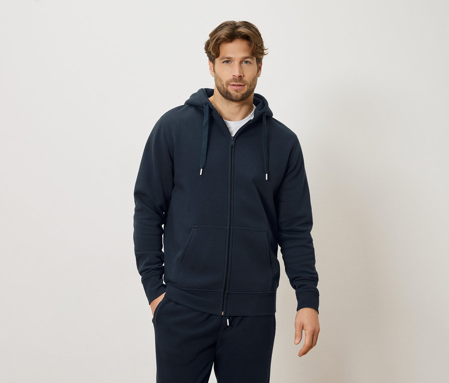 Férfi tengerészkék cipzáras kapucnis pulóverben és tengerészkék melegítőalsóban, krémszínű háttér előtt állva.