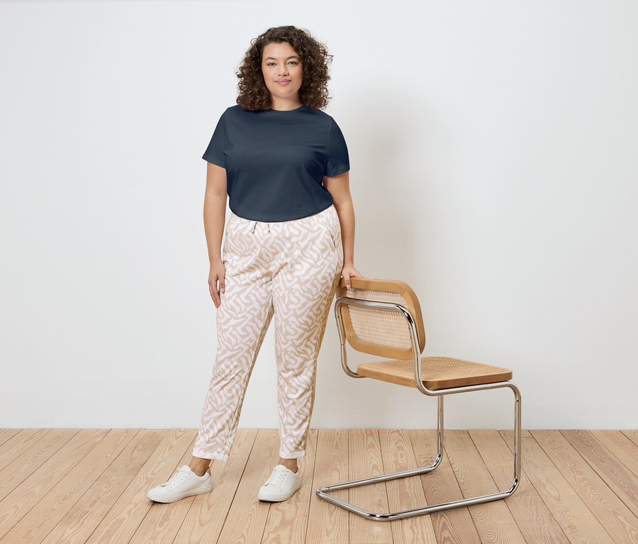 Egy göndör hajú nő áll egy fa keretes, rattan ülésű szék mellett. Sötétkék pólót, mintás nadrágot és fehér tornacipőt visel.