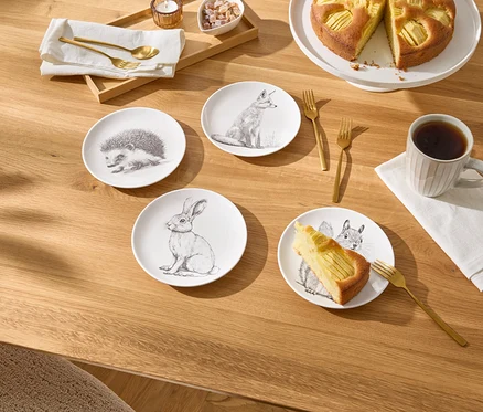 Egy fa asztalon négy kis tányér áll állatrajzokkal, egy szelet almás pite, egy csésze kávé és arany evőeszközök.
