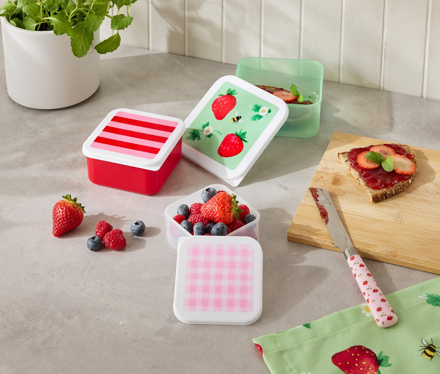 Számos színes ételtartó doboz áll a konyhapulton, némelyik gyümölcsökkel van megtöltve, egy szelet kenyér és egy kés mellett.