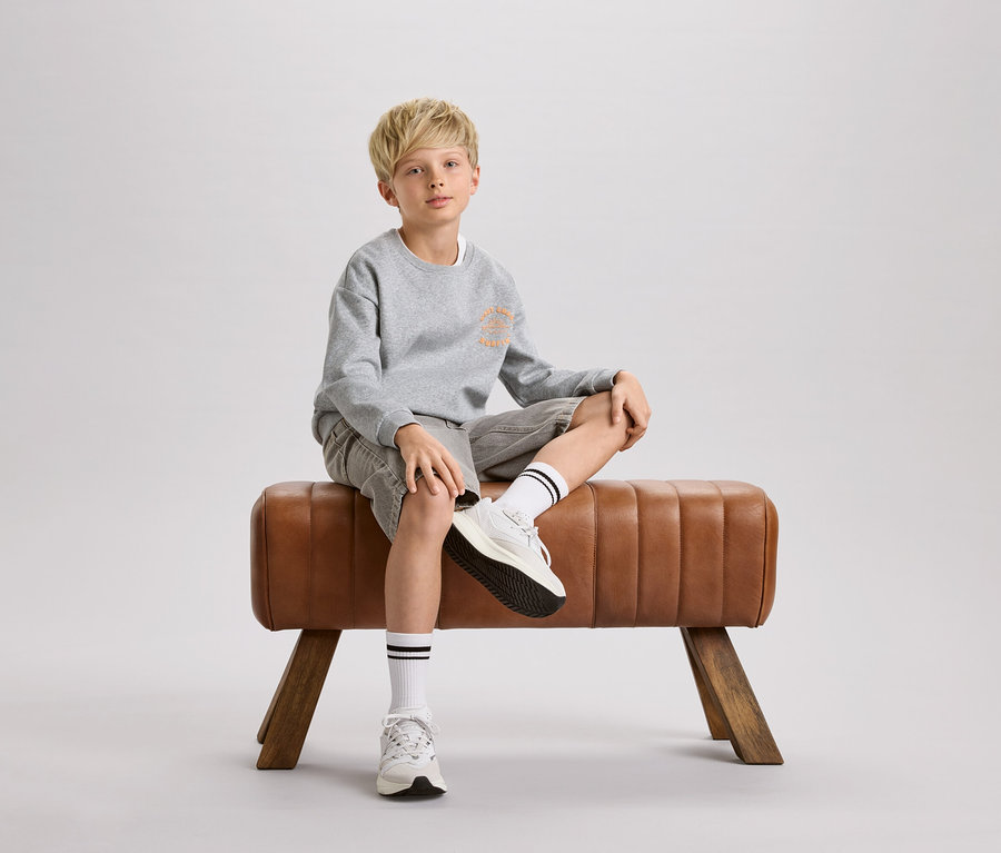 Egy szőke fiú ül egy barna tornabakon. Szürke pulóvert, rövidnadrágot és fekete csíkos fehér zoknit visel.