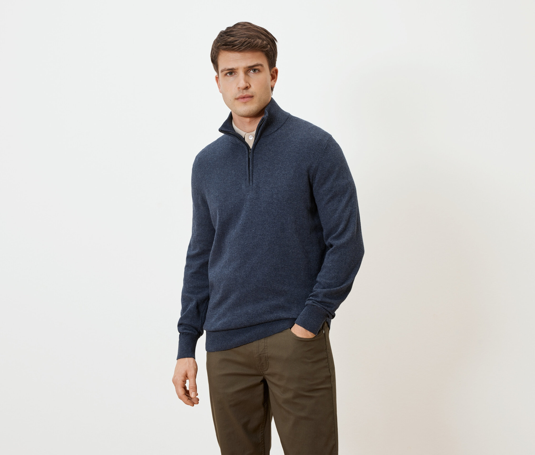 Férfi kék, cipzáras pulóverben és barna nadrágban, kezével a zsebében, semleges háttér előtt.