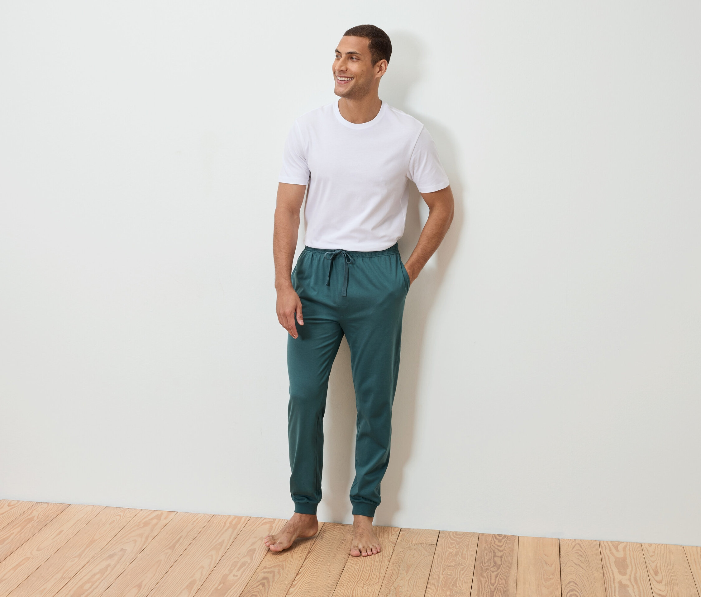 Egy férfi mezítláb áll, kezével a zsebében egy fehér fal előtt. Fehér pólót és petrolkék színű melegítőnadrágot visel.