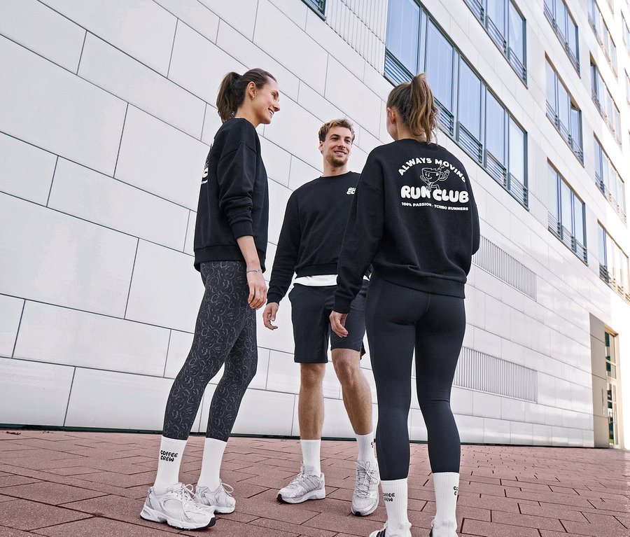 Három ember fekete szabadidőfelsőben és sportruházatban beszélget. Egy nő leopárdmintás, antracit női sportnadrágot és uniszex Coffe Crew teniszzoknit visel. Egy férfi fekete sport rövidnadrágot és fehér zoknit visel. Egy másik nő fekete női sportnadrágot és „RUN CLUB” logós szabadidőfelsőt visel.