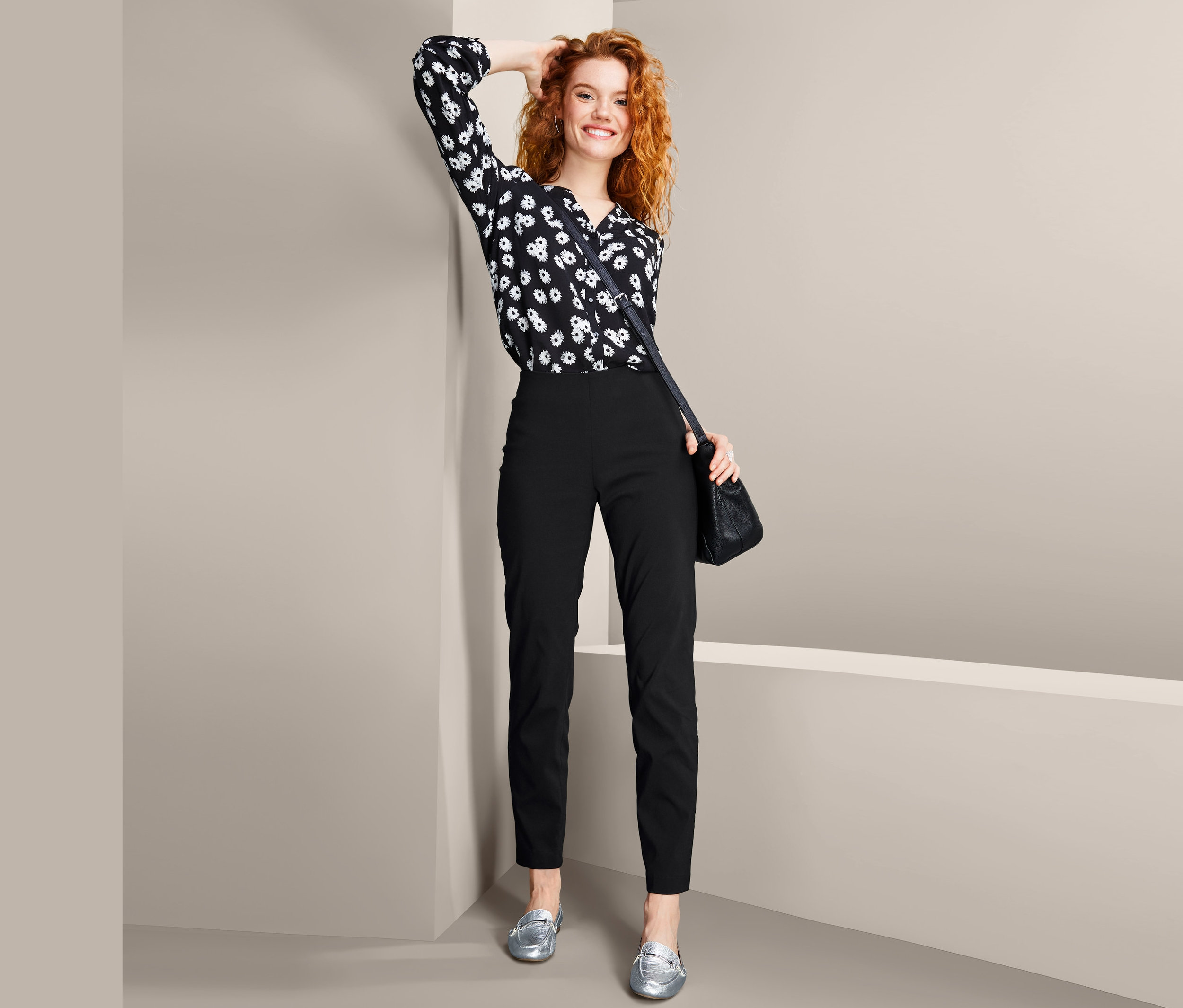Mosolygó nő pózol fekete, bokáig érő sztreccsnadrágban és mintás blúzban.
