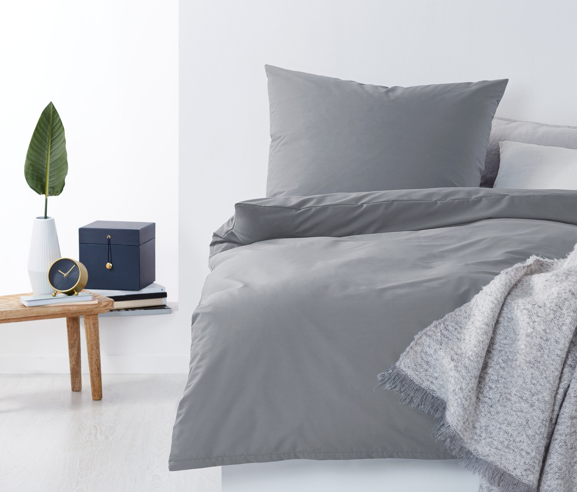 Szürke perkál ágyneműhuzat, egyszemélyes méretben egy ágyon, szürke takaróval. Éjjeliszekrény vázával és órával.