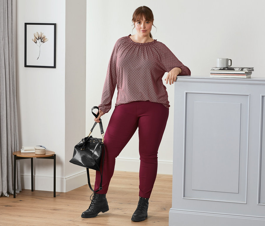 Nő pózol mintás, bordó női hosszú ujjú pólóblúzban, bordó sztreccsnadrágban, fekete női bőrbakancsban és fekete, krokodilbőr hatású női válltáskával.