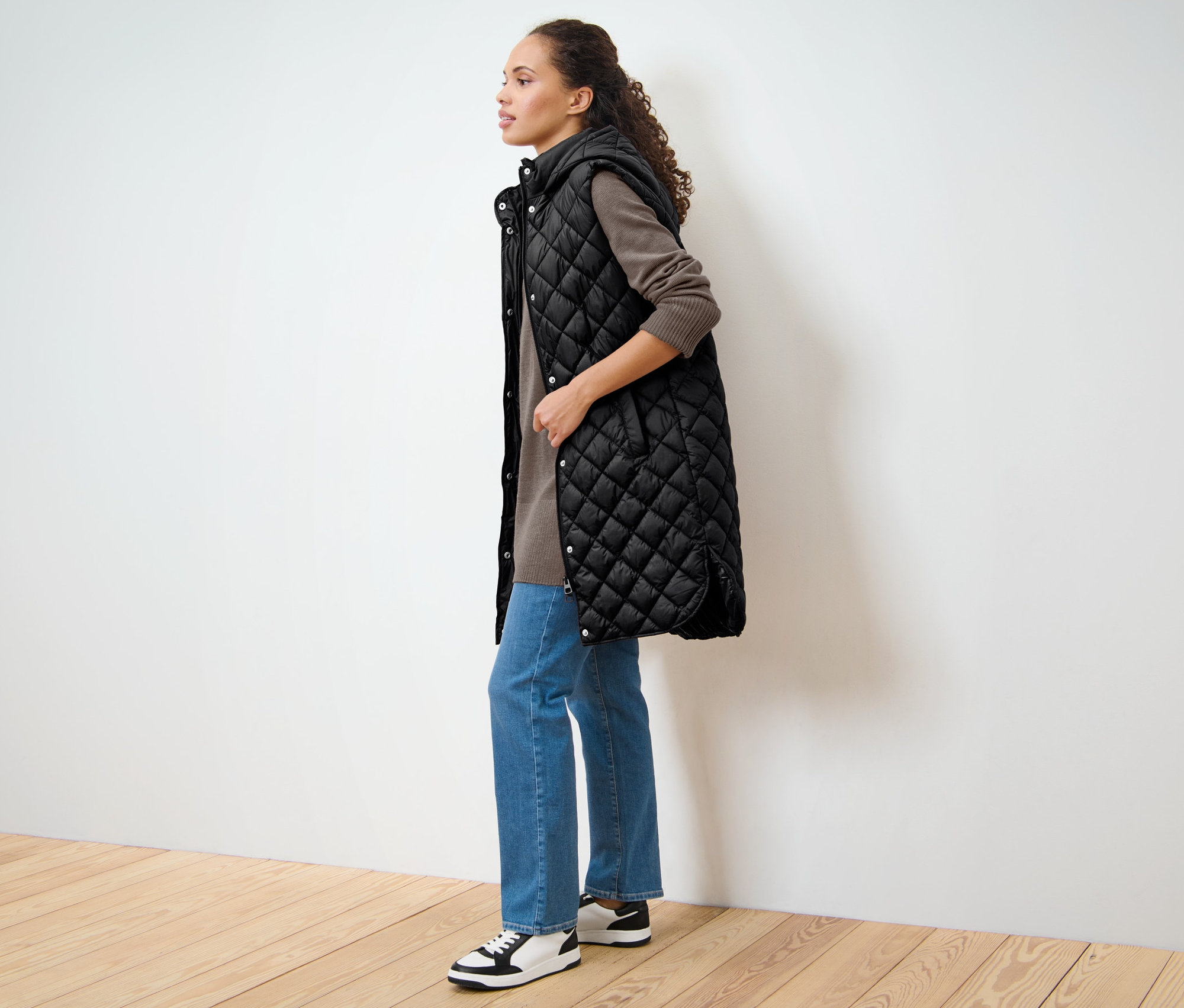 Nő áll oldalra, és egy fekete, hosszú steppelt mellényt visel.