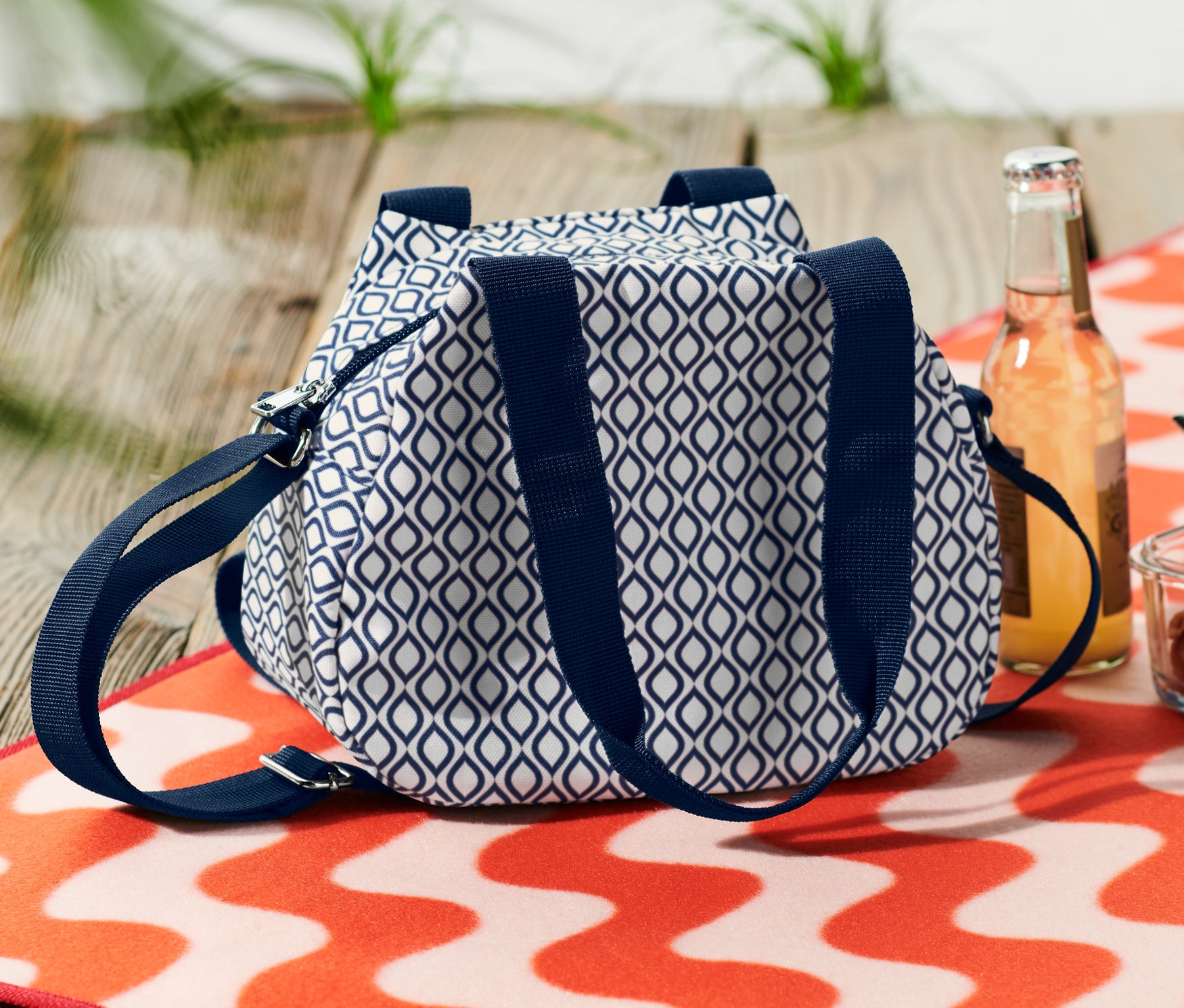 Egy kék-fehér mini hűtőtáska egy ital és egy tésztás tál mellett fekszik egy narancs-fehér hullámos strandtörölközőn.
