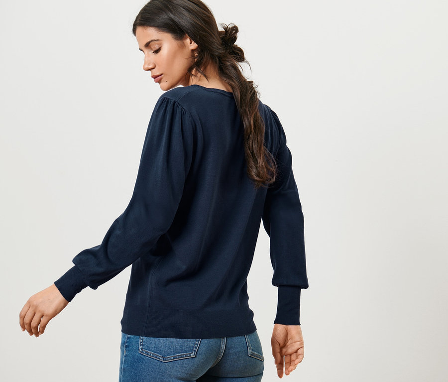 Nő pózol egy női finomkötésű pulóverben és farmerben.