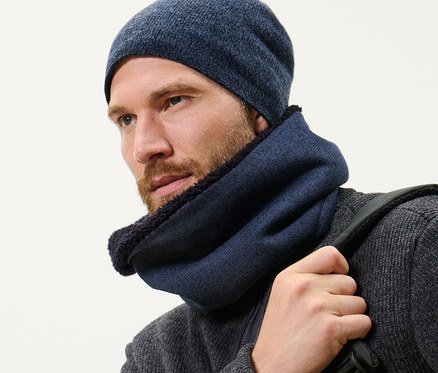 Szakállas férfi sötétkék kötött polár csősálat és sapkát visel, hátizsákot visz.