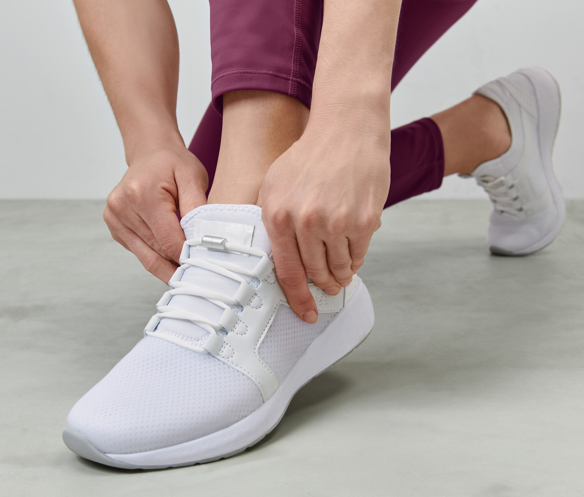 Nő fűzi az elasztikus cipőfűzőt egy fehér cipőn.
