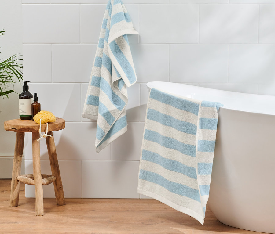 Két világoskék-fehér csíkos törölköző lóg a fürdőszobában egy fürdőkád mellett.